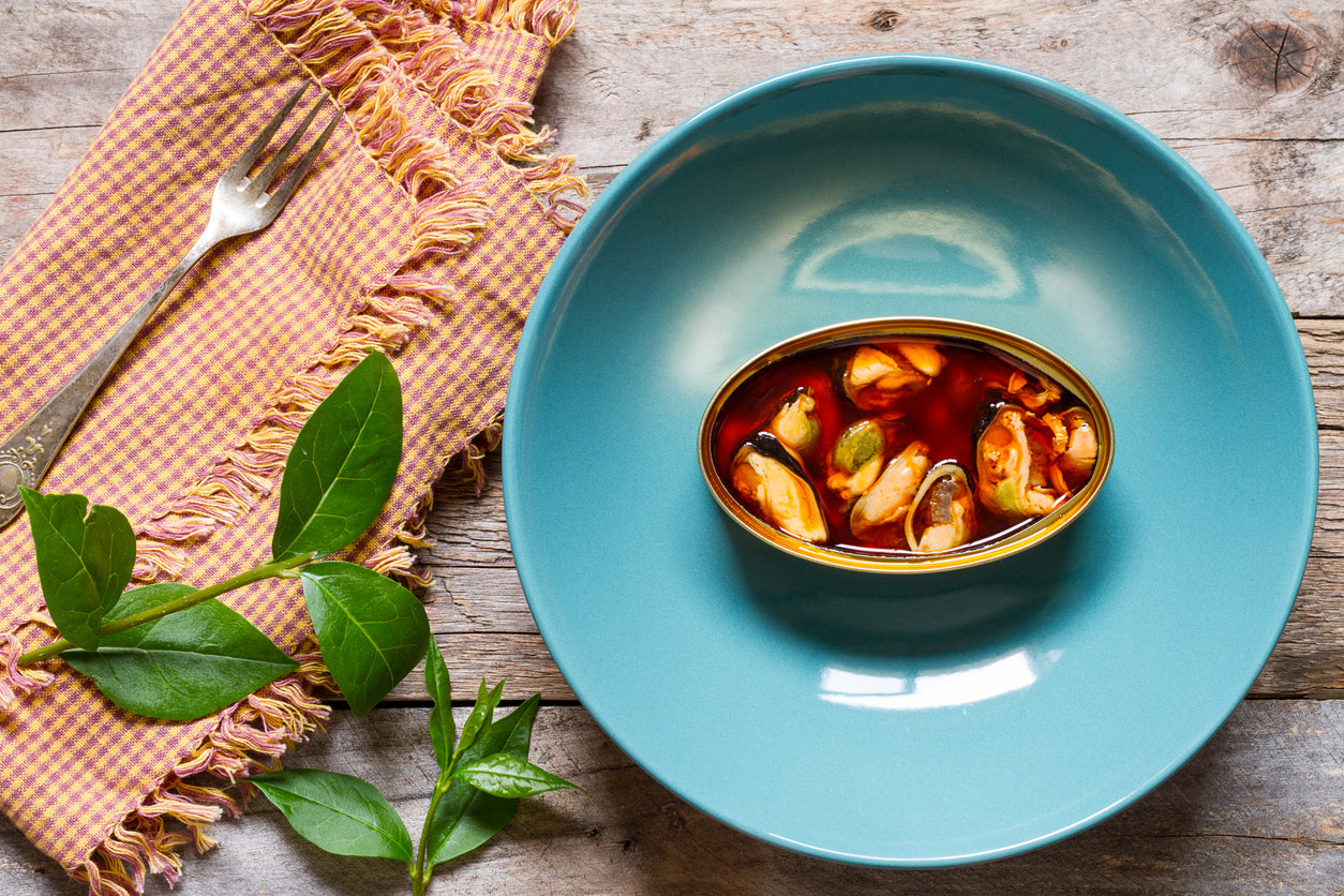 a turquoise plate containing an open oval can of tinned mussels in a red oil sauce. A napkin with a fork and a green branch are to the left of the plate