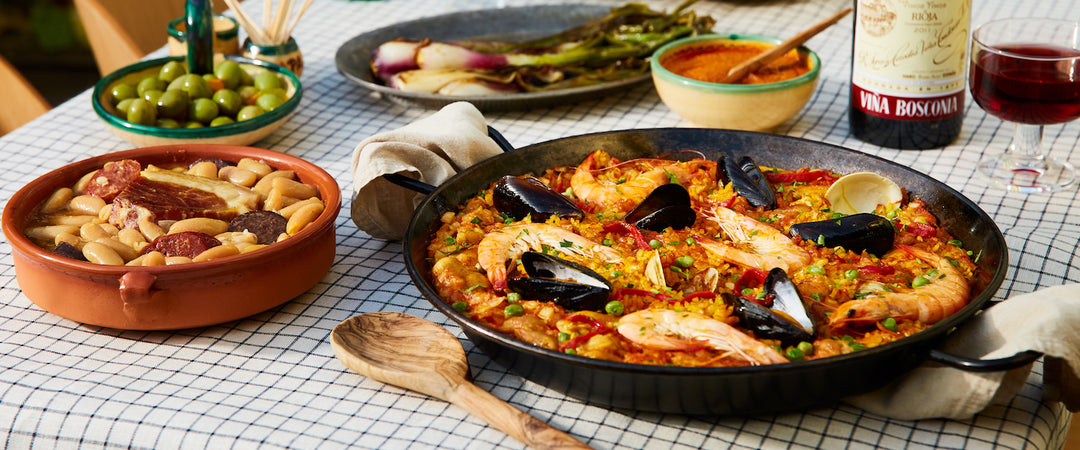 a table set with a freshly cooked seafood paella in a paella pan surrounded by cooked beans, a dish with green olives, some grilled green onions and a bottle of red wine with an open glass next to it.